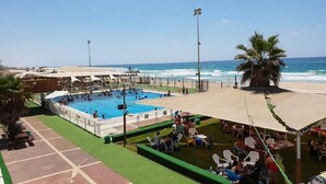 A pool area with a view of the ocean, surrounded by a sandy beach and palm trees. - Masarik Apartments (Bat Yam)