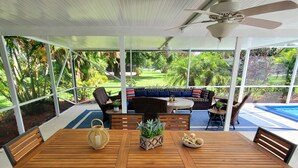 Un patio couvert avec une table et des chaises en bois, un ventilateur de plafond et une vue sur une piscine et un jardin.