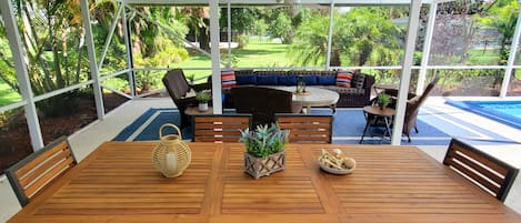 Un patio couvert avec une table et des chaises en bois, un ventilateur de plafond et une vue sur une piscine et un jardin.