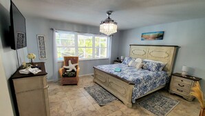 Une chambre à coucher comprenant un lit, une chaise, une commode, une table de chevet, un téléviseur et une fenêtre avec des stores.