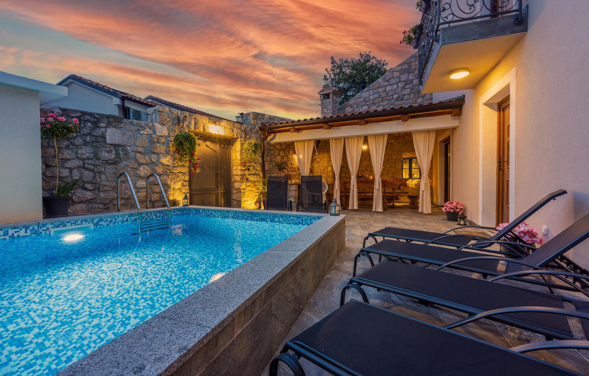 A swimming pool with lounge chairs, stone walls, and a sunset sky.