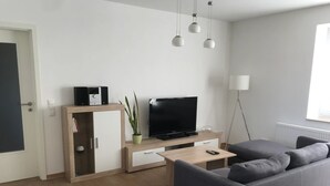 A modern living room with a grey sofa, a wooden coffee table, a TV on a stand, a potted plant, and a floor lamp. - Vacation apartment in the heart of Saxony (Leisnig)