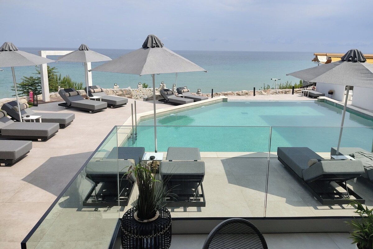 A pool area with lounge chairs and umbrellas, overlooking the sea.