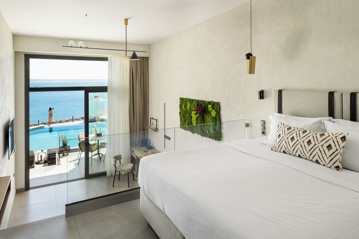Una habitación de hotel moderna con una cama grande, un balcón de cristal con vista al mar, un escritorio pequeño y un adorno de pared verde.