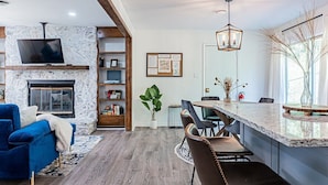 A modern living room with a stone fireplace, a flat-screen TV, a blue sofa, and a dining area with a marble countertop and bar stools. - Modern 3BR Family Home, Fenced Yard, Pets Friendly + 15 min to Stone Mountain (Snellville)