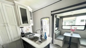 A compact kitchenette with a sink, stove, and cabinets. There is a small table with a vase of flowers and a window with blinds. - Johnny Love, Modern and Bright Paradise RV (San Diego)