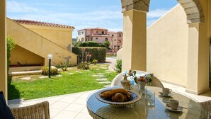 Un espace patio avec une table ronde en verre, des chaises en osier, un panier de fruits et une vue sur un jardin et des bâtiments en arrière-plan.