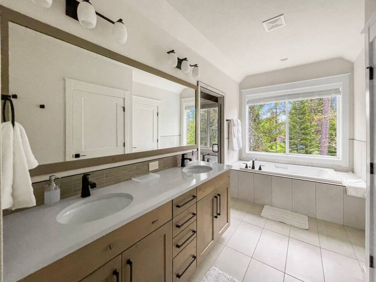 A modern bathroom with two sinks, a large mirror, and a window providing a view of trees.