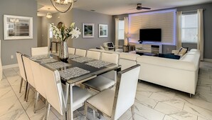 A modern living room with a dining area, white furniture, a flat-screen TV, and artwork on the walls.