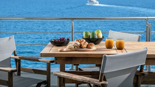 Taula de fusta amb preparació per a l'esmorzar, incloent fruita, pastissos i suc, amb vista a un mar tranquil on es pot veure un vaixell a la distància.