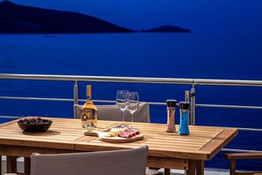 Una mesa dispuesta con una botella de vino, dos copas, un cuenco de cerezas, una tabla de quesos y dos saleros y pimenteros sobre una mesa de madera con vistas al mar y las montañas al fondo.
