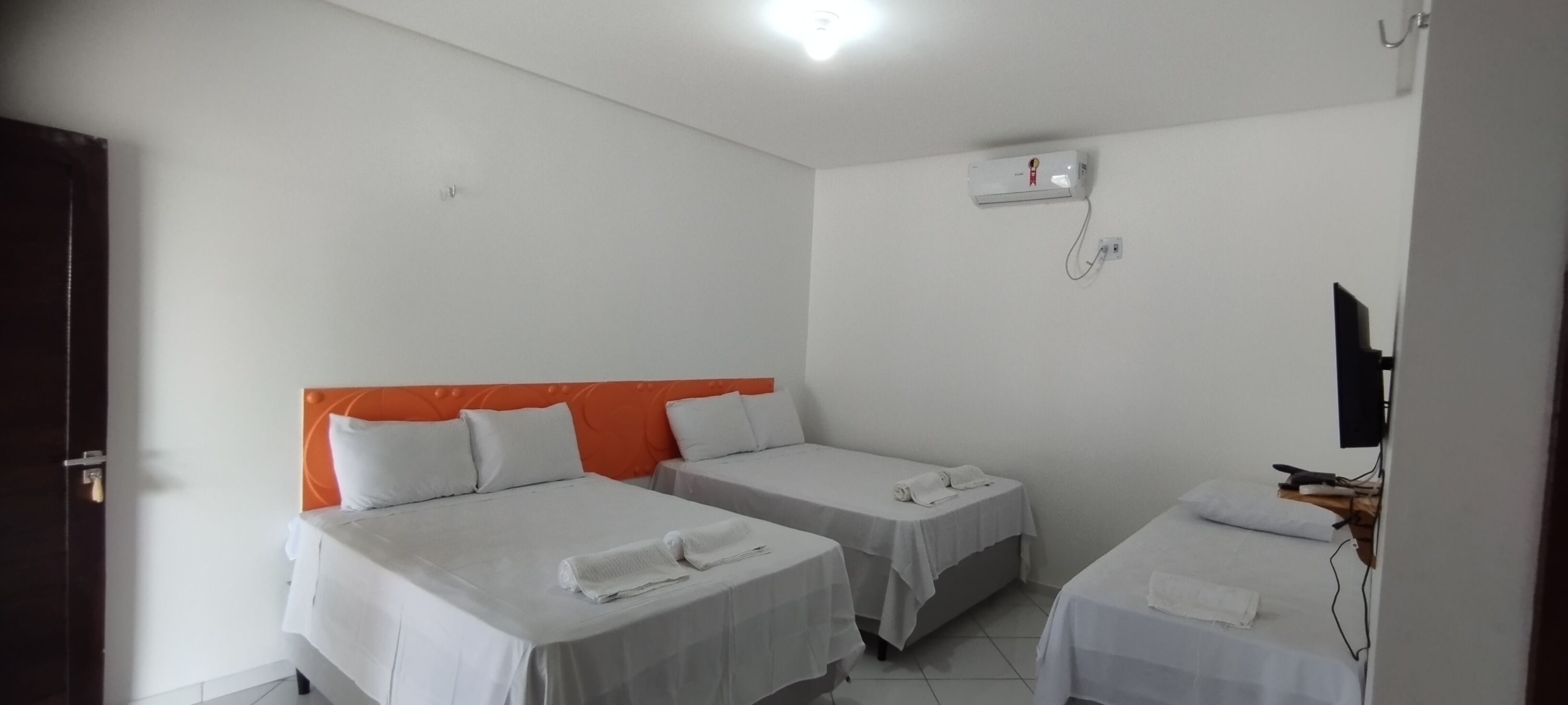 Ein Hotelzimmer mit zwei Einzelbetten, einer weißen Deckenleuchte, einer Wand-Klimaanlage und einem Fernseher auf einem Holzboden.