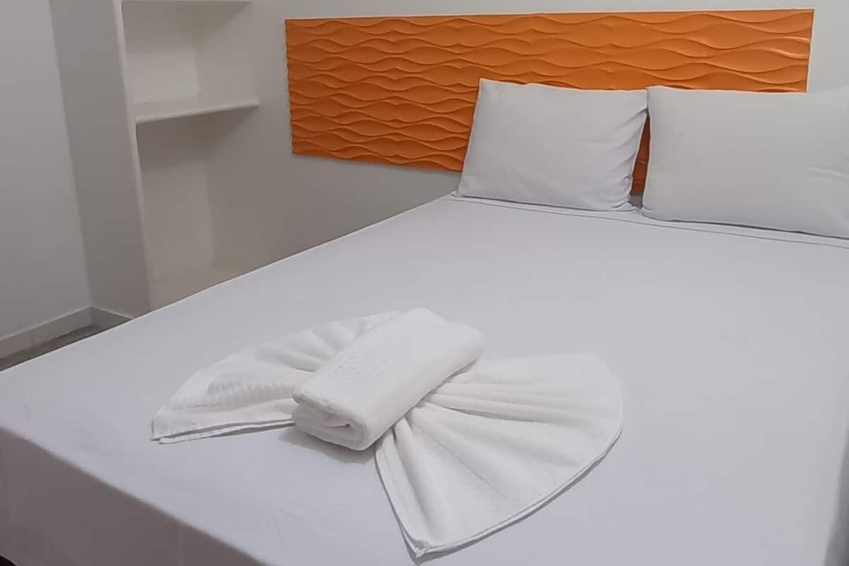 A hotel room with a bed, white walls, a wooden headboard, and a white bedspread.