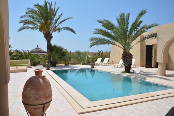 Relax by the serene pool surrounded by palm trees and a thatched-roof structure.