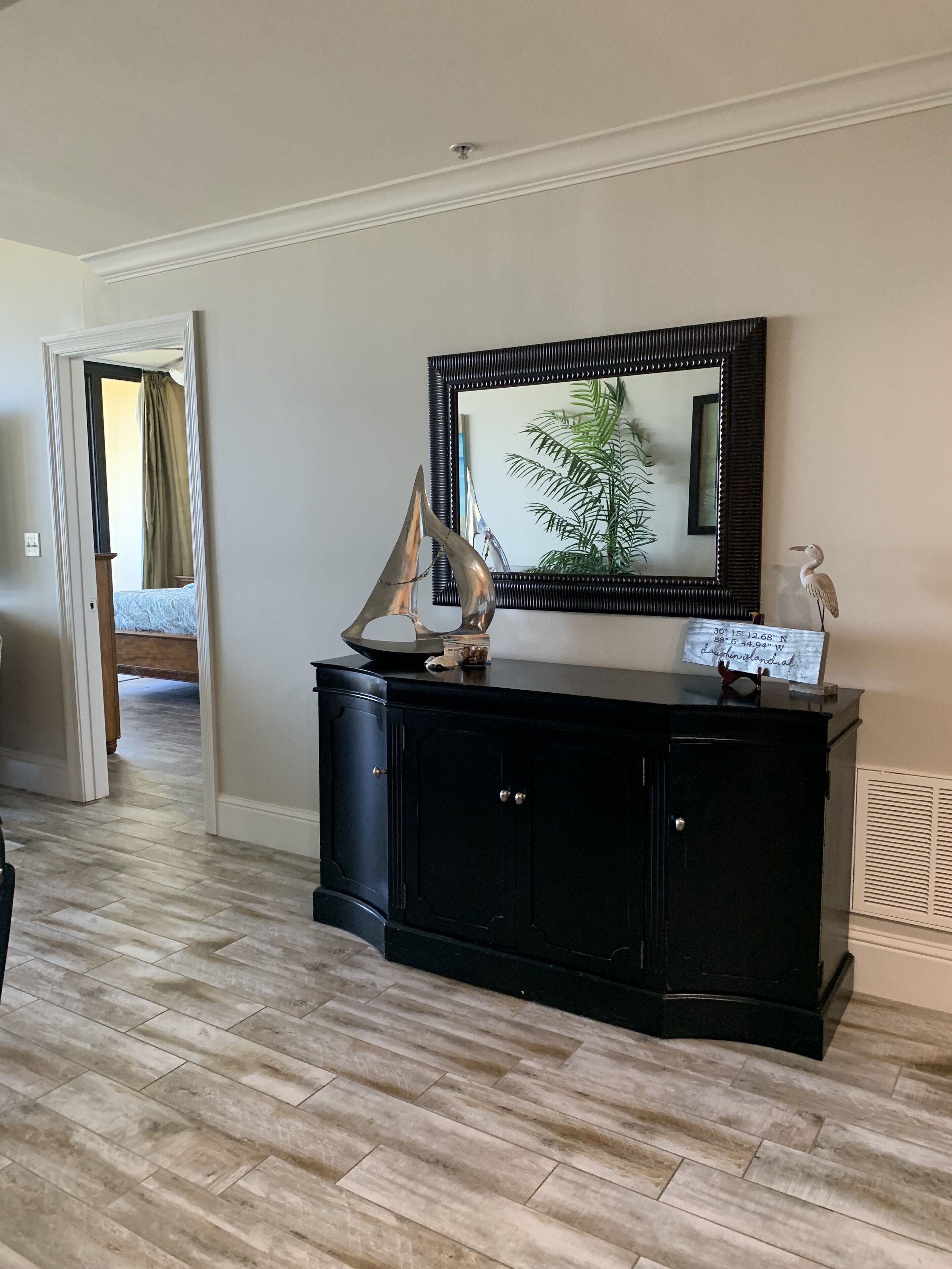 A room with a black console table, a large mirror, and a framed picture of a plant.