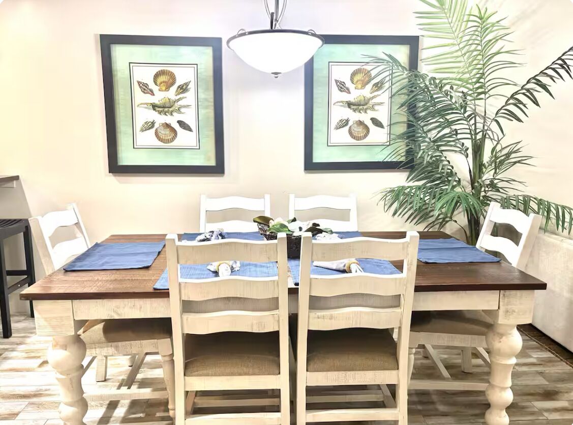 Un coin repas avec une table en bois et quatre chaises, des œuvres d’art encadrées au mur et une grande plante en pot.