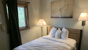 A neatly made bed with white bedding, two bedside lamps, a window with curtains, and a painting of a tree on the wall.