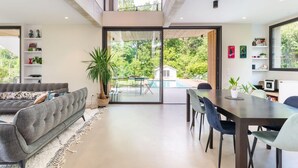 En moderne stue med en grå sofa, et spisebord med stole, et stort vindue med udsigt til pool og grønt område, samt en trappe til en etage over.