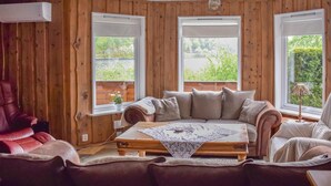 Ein gemütliches Blockhäuschen mit Holzvertäfelung, einem Ledersofa, einem Holztisch und großen Fenstern mit Blick auf Grünflächen.