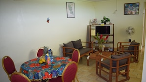 A dining area with a round table, red chairs, and a floral tablecloth. There is a sofa and a TV stand with a plant and a fan.