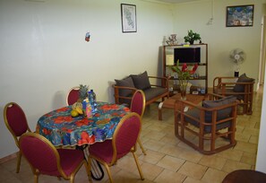 Ein Essbereich mit einem runden Tisch, roten Stühlen und einer Tischdecke mit Blumenmuster. Es gibt ein Sofa und eine TV-Konsole mit einer Pflanze und einem Ventilator.
