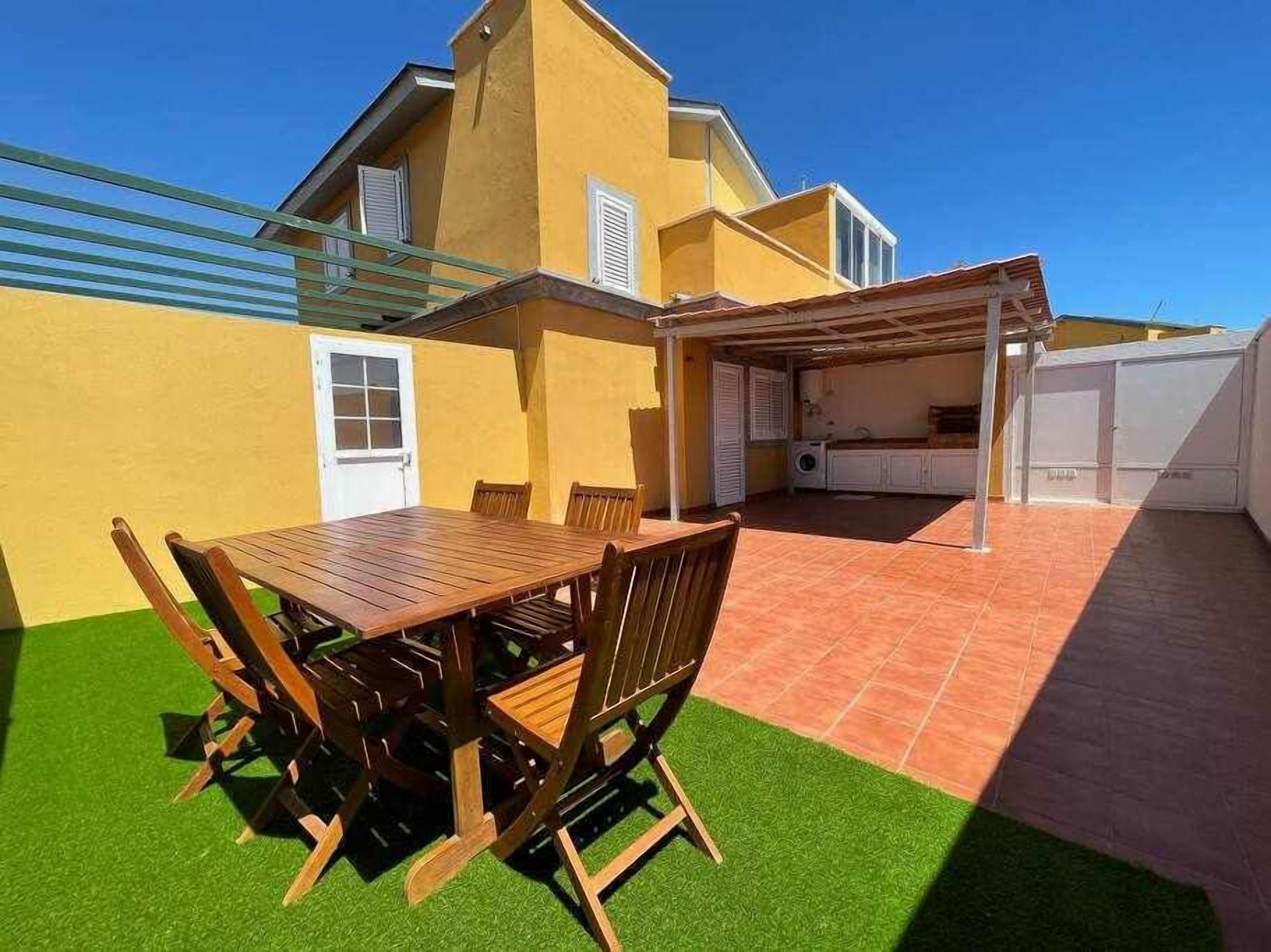 A patio with a wooden table and chairs, a covered seating area, and a building with a yellow exterior and white doors.