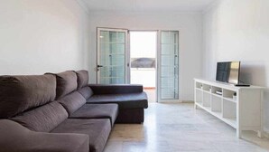 Ein modernes Wohnzimmer mit einem Ecksofa, einem Fernseher auf einer weißen Konsole und einer Glastür, die nach draußen führt.