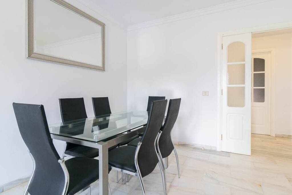 Une salle de conférence avec une table en verre et quatre chaises noires, un grand miroir au mur et une porte blanche avec un panneau en verre.