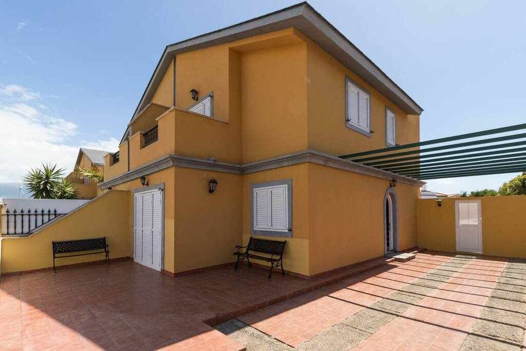 Un edificio de dos pisos con fachada amarilla, un patio cubierto y un pequeño jardín.