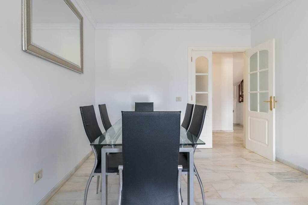 A dining area with a glass table and four chairs, a mirror on the wall, and a white door leading to another room.