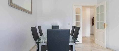 A dining area with a glass table and four chairs, a mirror on the wall, and a white door leading to another room.