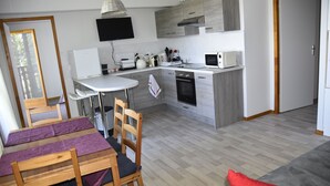 A modern kitchen with white appliances, a dining area with wooden chairs and a purple tablecloth, and a grey sofa with a red pillow.