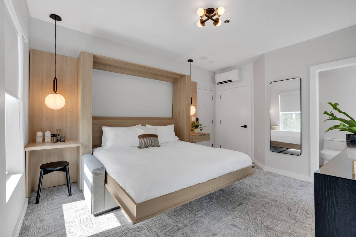 A modern bedroom with a wooden headboard, a large bed, bedside tables, a desk, and a mirror.