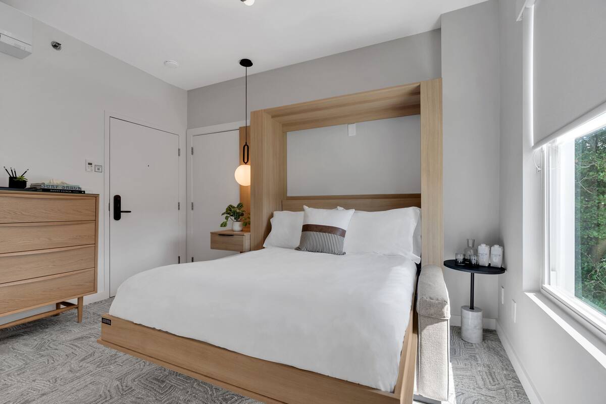 A modern bedroom with a wooden bed, a bedside table with a lamp, and a view of greenery through the window.