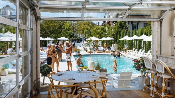 Un área de piscina al aire libre con sillas de descanso, sombrillas y un porche con paredes de vidrio.