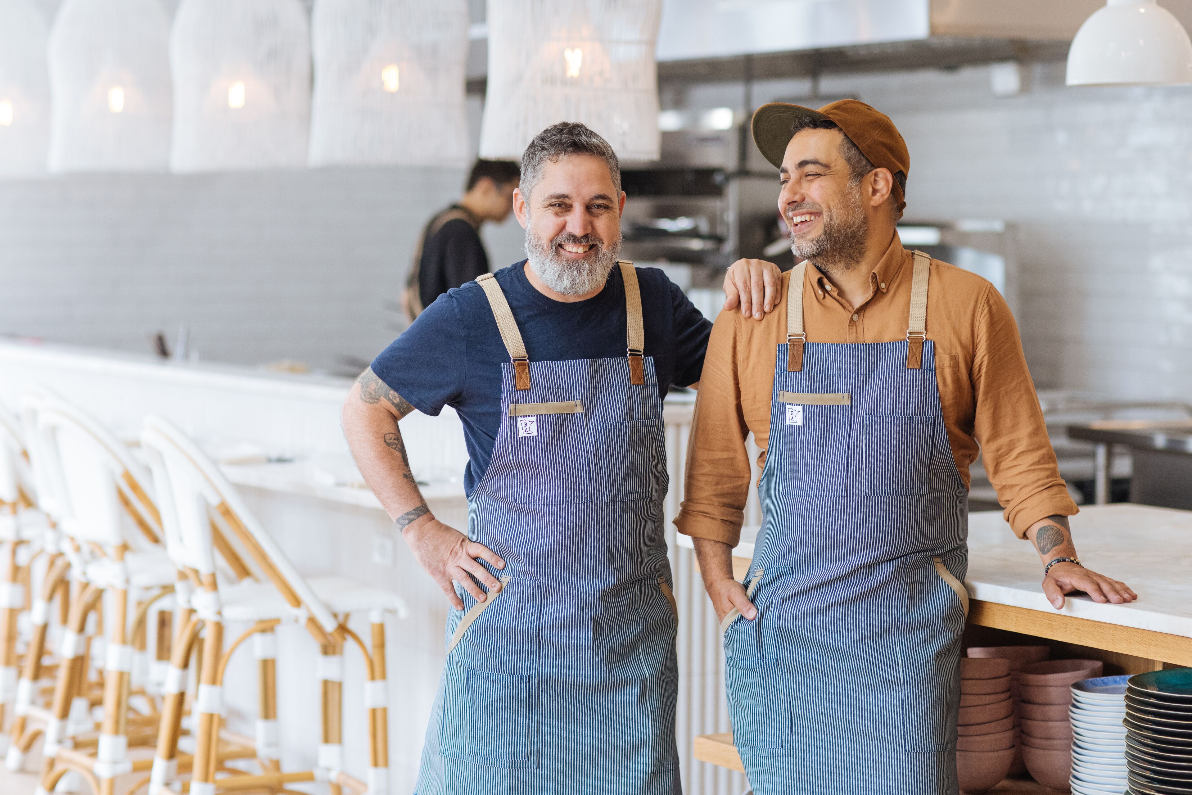 Two individuals in aprons standing in a kitchen area with white chairs and a counter.