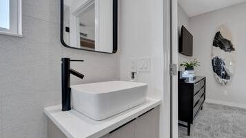 A modern bathroom with a white sink, black faucet, and a mirror above it. There is a black dresser with a plant on top and a surfboard decoration on the wall.