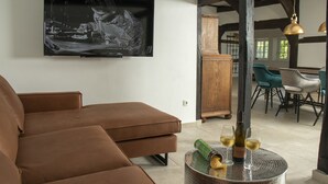 A modern living room with a brown sofa, a round coffee table with a bottle and glasses, and a flat-screen TV on the wall. - Dream cottage (Menslage)