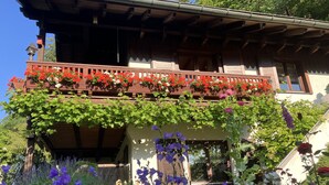 A traditional wooden house with a balcony adorned with red and white flowers, and a garden with purple flowers in front. - Ferienwohnung mit Fantastischem Ausblick, Direkt am Wald (Walkenried)