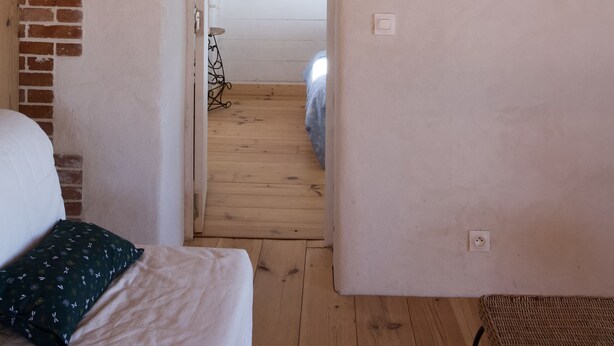 Een smalle gang met een bed aan de linkerkant, een houten vloer en een deur naar een andere kamer.