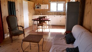 Een gezellig houten interieur met vloer, een eethoek met stoelen en tafel, een bank met kussens, een keuken met een magnetron en kastjes, en een groot raam.