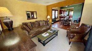 Un salon avec un canapé, une table basse et un fauteuil en osier. On aperçoit une lampe sur une table, un miroir au mur et, au fond, un coin cuisine.