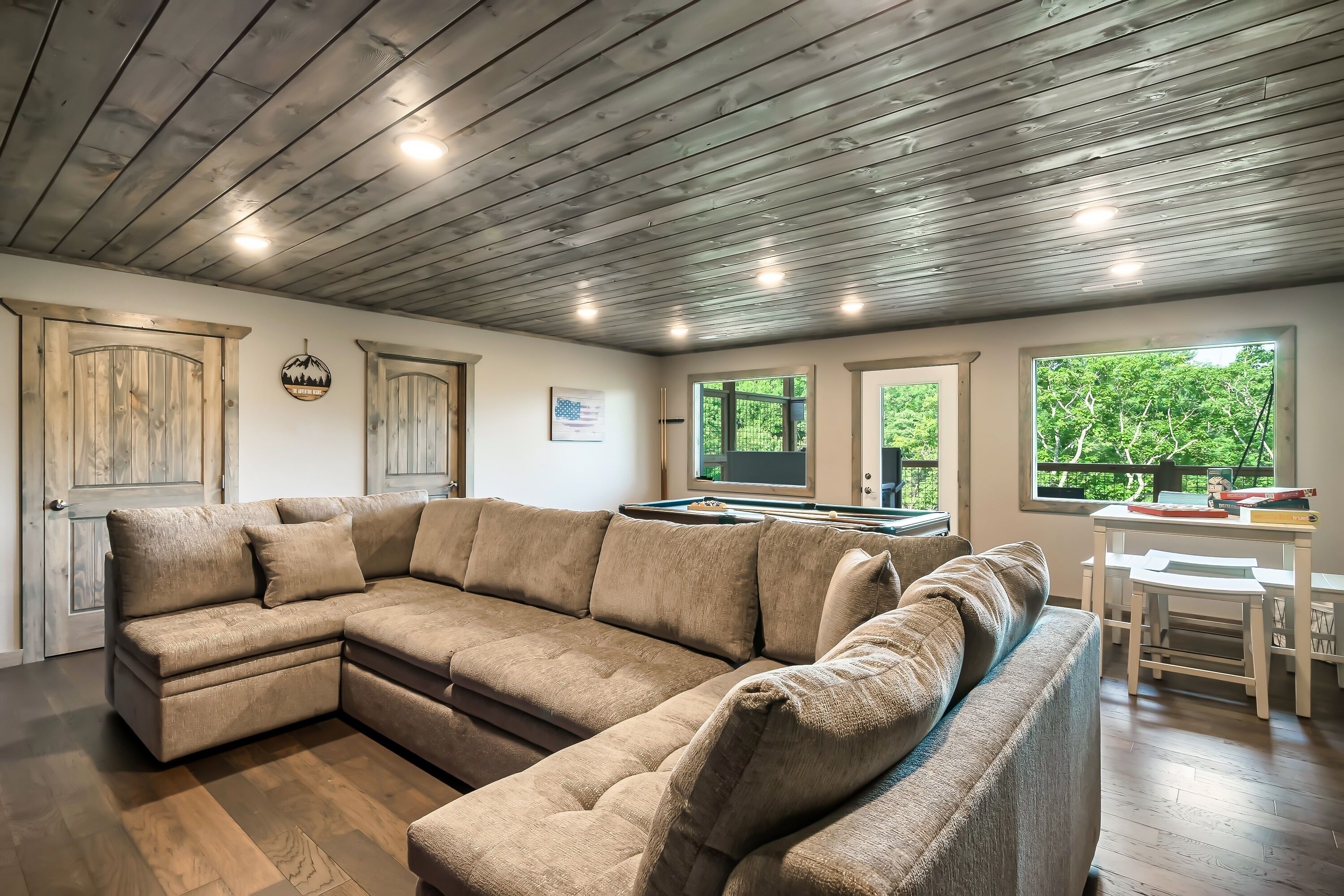 A modern living room with a sectional sofa, wooden ceiling, and large windows offering a view of greenery.