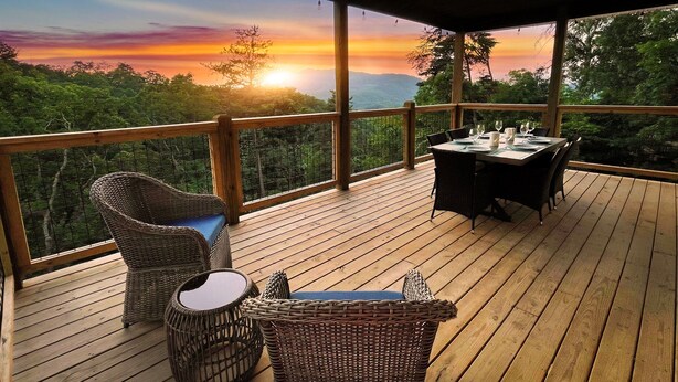 Une terrasse en bois, agrémentée de meubles en osier et d’une table dressée pour le dîner, offrant une vue sur un paysage boisé au coucher du soleil.