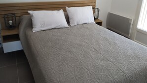 A bedroom with a bed, two white pillows, a wooden headboard, a nightstand with a lamp, and a radiator. - Large air-conditioned T2, private parking near the center and beach (Banyuls-sur-Mer)