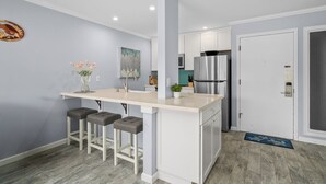 A modern kitchen with a central island, bar stools, a refrigerator, and a door with a keypad lock. - Plantation Hale J2 | Relaxing Kapaa Escape on Kauai's Coconut Coast (Kapaa)