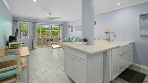 A modern kitchen with a central island, a dining area with a table and chairs, and a view through the glass doors. - Plantation Hale J2 | Relaxing Kapaa Escape on Kauai's Coconut Coast (Kapaa)