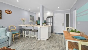 A modern kitchen with white cabinets, a central island, and a dining area with a wooden table and chairs. - Plantation Hale J2 | Relaxing Kapaa Escape on Kauai's Coconut Coast (Kapaa)
