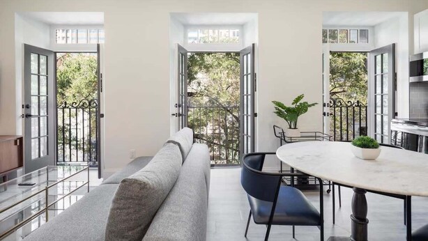 Un salon moderne avec un canapé, une table ronde et des chaises. Des portes ouvertes donnent sur un balcon d’où l’on peut admirer la vue sur les arbres.