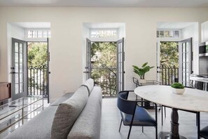 Un salon moderne avec un canapé, une table ronde et des chaises. Des portes ouvertes donnent sur un balcon d’où l’on peut admirer la vue sur les arbres.